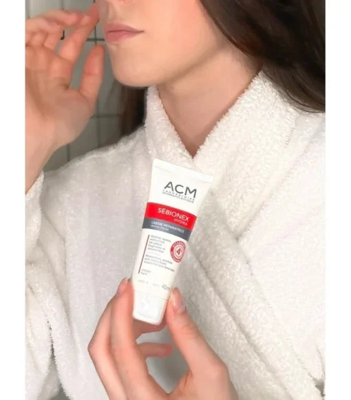 Femme en peignoir appliquant la crème réparatrice ACM Sébionex Hydra sur son visage.