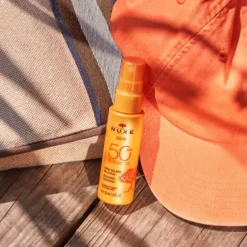 Spray solaire SPF 50 NUXE Sun posé à côté d'une casquette orange
